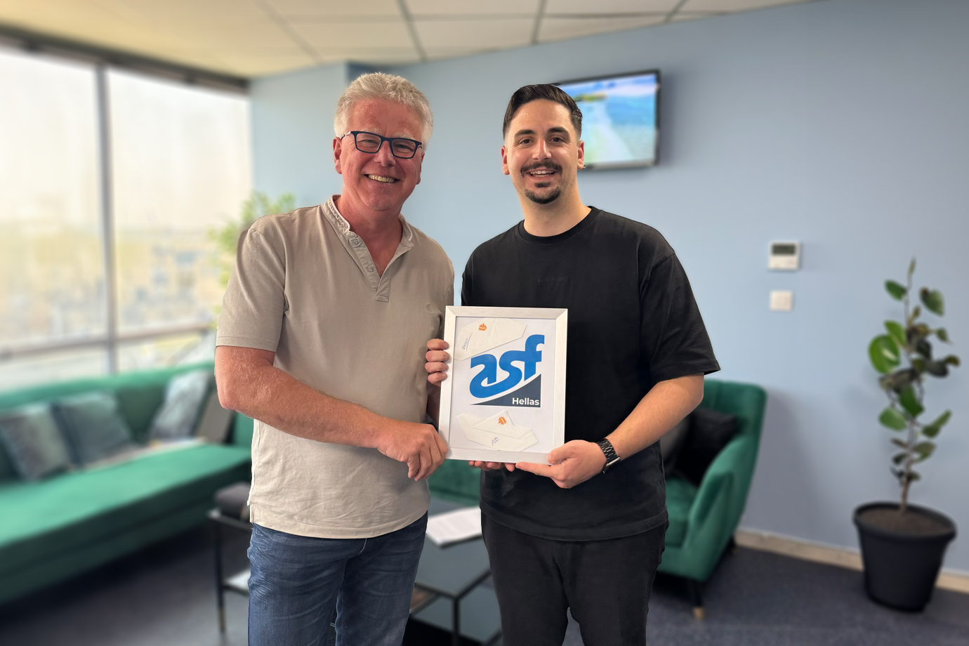 Martin Aye, Inhaber der aye media marketing group, überreicht Ioannis Gisgakis, Geschäftsführer der asf hellas, einen Bilderrahmen, der das Logo der asf hellas enthält. Im Hintergrund sind eine Fensterfront und ein grünes Sofa zu sehen.